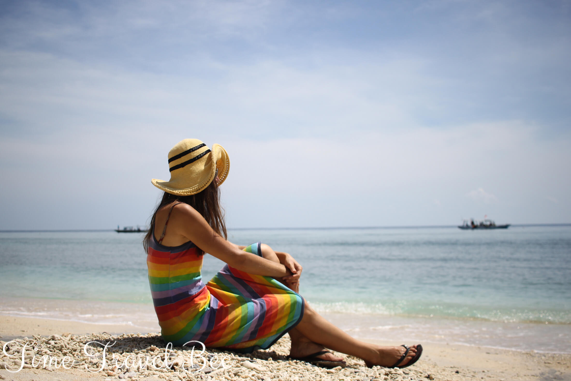 Dziewczyna w tęczowej sukience i żółtym kapeluszu. Plaża na wyspie Gili, Lombok. Statki w tle i turkusowa woda.